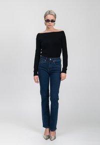 Top lungo a maniche lunghe nera off-the-shoulder, jeans blu a vita alta e scarpe con punta a righe. Modella su uno sfondo neutro.