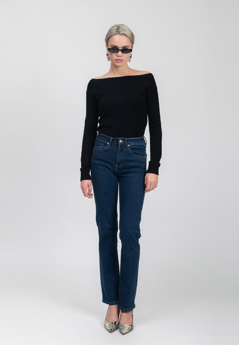 Top lungo a maniche lunghe nera off-the-shoulder, jeans blu a vita alta e scarpe con punta a righe. Modella su uno sfondo neutro.