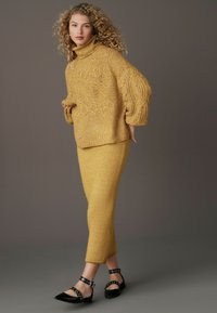 Mosterdkleurige gebreide coltrui met textuurpatronen, gecombineerd met een aansluitende, glinsterende mosterdkleurige rok en zwarte puntige flats met studs.
