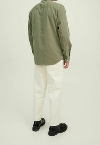 Camicetta verde oliva a maniche lunghe con colletto button-down, abbinata a pantaloni bianchi e scarpe nere, vista da dietro.