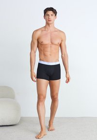 Boxeri negri cu un brâu elastic alb. Modelul stă desculț, evidențiind un design ajustat și o textură a țesăturii netedă.