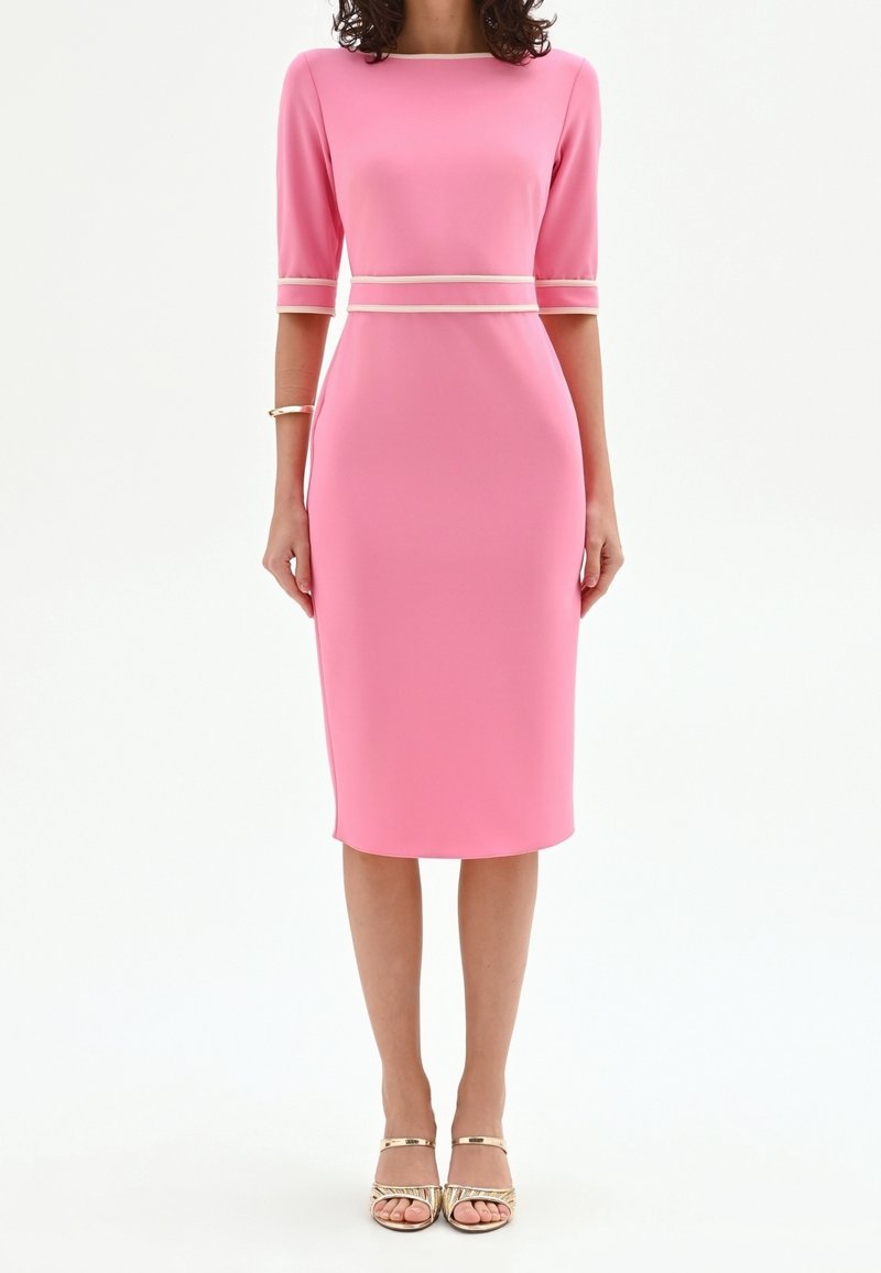 Femme portant une robe midi rose ajustée avec des manches trois-quarts et une bordure claire à la taille et aux poignets, associée à des talons ouverts dorés.