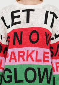 Pullover mit breiten Streifen in Weiß, Rot, Pink und Grün, der in fettem Schwarz und Rot den Text "LASS ES SCHNEIEN, FUNKELN, GLÜHEN" zeigt.