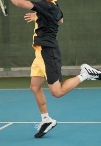 Czarny i żółty strój sportowy z krótkim rękawem i szortami. Czarnobiałe sneakersy. Niebieska nawierzchnia kortu tenisowego w tle.