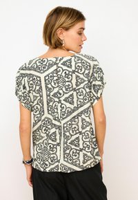 T-shirt z krótkim rękawem w kolorze kremowym z czarnym geometrycznym wzorem, z wycięciem w kształcie klucza z tyłu i luźnym krojem. Miękka struktura.
