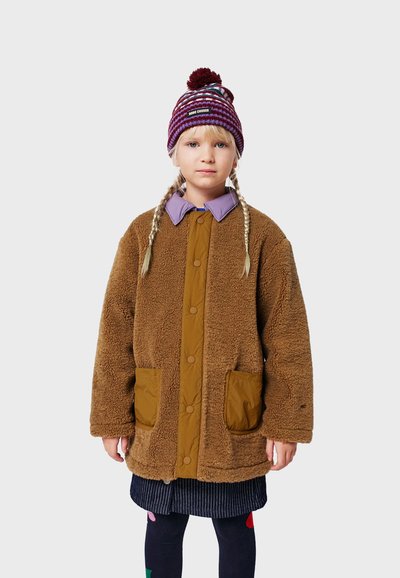 Braune Fleecejacke mit einem sandfarbenen Vorderteil, ausgestattet mit zwei Vorderfächern. Kombiniert mit einem lila Hemd mit Kragen und einer gemusterten Mütze.