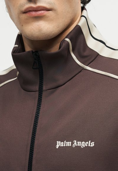 Κλείσιμο ενός ατόμου που φοράει καφέ ζακέτα με φερμουάρ με κρεμ ρίγες και το κείμενο "Palm Angels" στο στήθος.