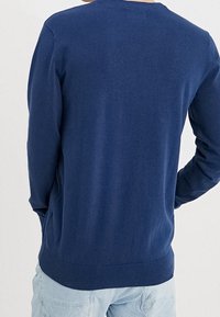 Uomo vestito con un maglione a maniche lunghe blu navy semplice e jeans azzurri, visto da dietro.