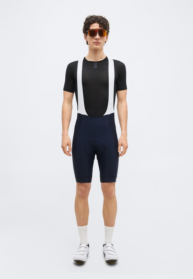 Young man in black cycling bib shorts, short-sleeve black jersey, white socks, white cycling shoes, and orange-tinted sports sunglasses.