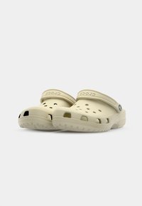 Sabots Crocs de couleur sable, fabriqués en matériau léger, dotés de trous de ventilation, d'une sangle arrière, d'une semelle texturée et d'un logo de marque en relief.