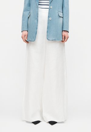 Person wearing white wide-leg pants, a light blue denim blazer over a striped shirt, and black pointed shoes standing still.