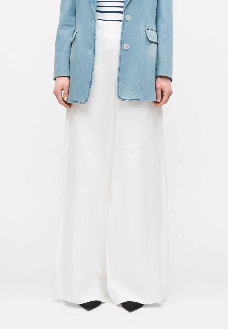 Άτομο που φοράει άσπρο παντελόνι φαρδιάς γραμμής, ένα ανοιχτό μπλε τζιν σακάκι πάνω από μια ριγέ μπλούζα και μαύρα μυτερά παπούτσια, στέκεται ακίνητο.