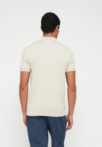 Polo bege de mangas curtas, com colarinho clássico e textura subtil. Usada com calças azul-marinho, vista mostra o design traseiro.