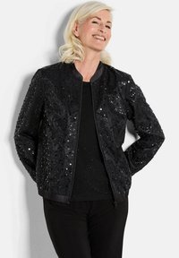 Veste bomber noire ornée de sequins, avec un col rond et des poignets côtelés. Portée sur un haut noir, associée à un pantalon noir.
