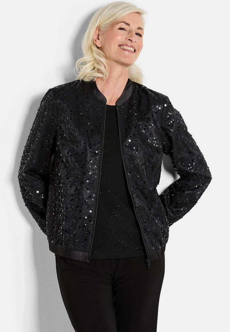 Veste bomber noire ornée de sequins, avec un col rond et des poignets côtelés. Portée sur un haut noir, associée à un pantalon noir.