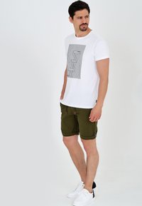 T-shirt en coton blanc avec un motif géométrique noir, associé à un short vert olive avec des ourlets roulés et un cordon de serrage. Baskets blanches.