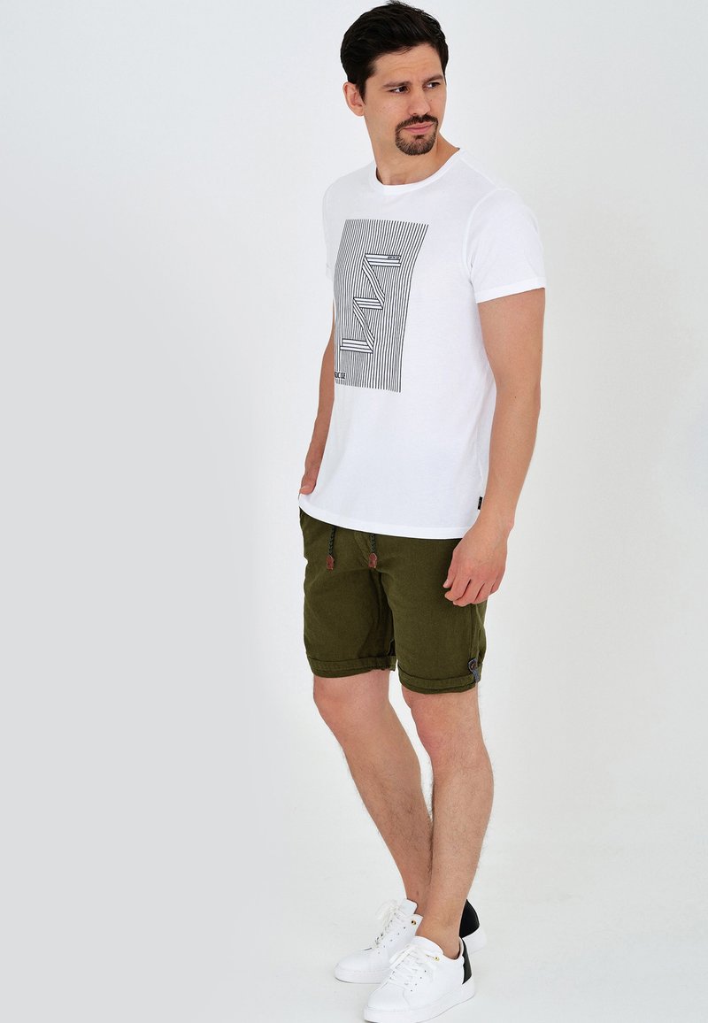 T-shirt en coton blanc avec un motif géométrique noir, associé à un short vert olive avec des ourlets roulés et un cordon de serrage. Baskets blanches.