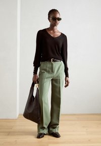 Schwarzer langärmliger V-Ausschnitt-Pullover, grüne weite Hose, schwarze Tragetasche mit goldenen Beschlägen und schwarze Slipper mit Kettenakzent.