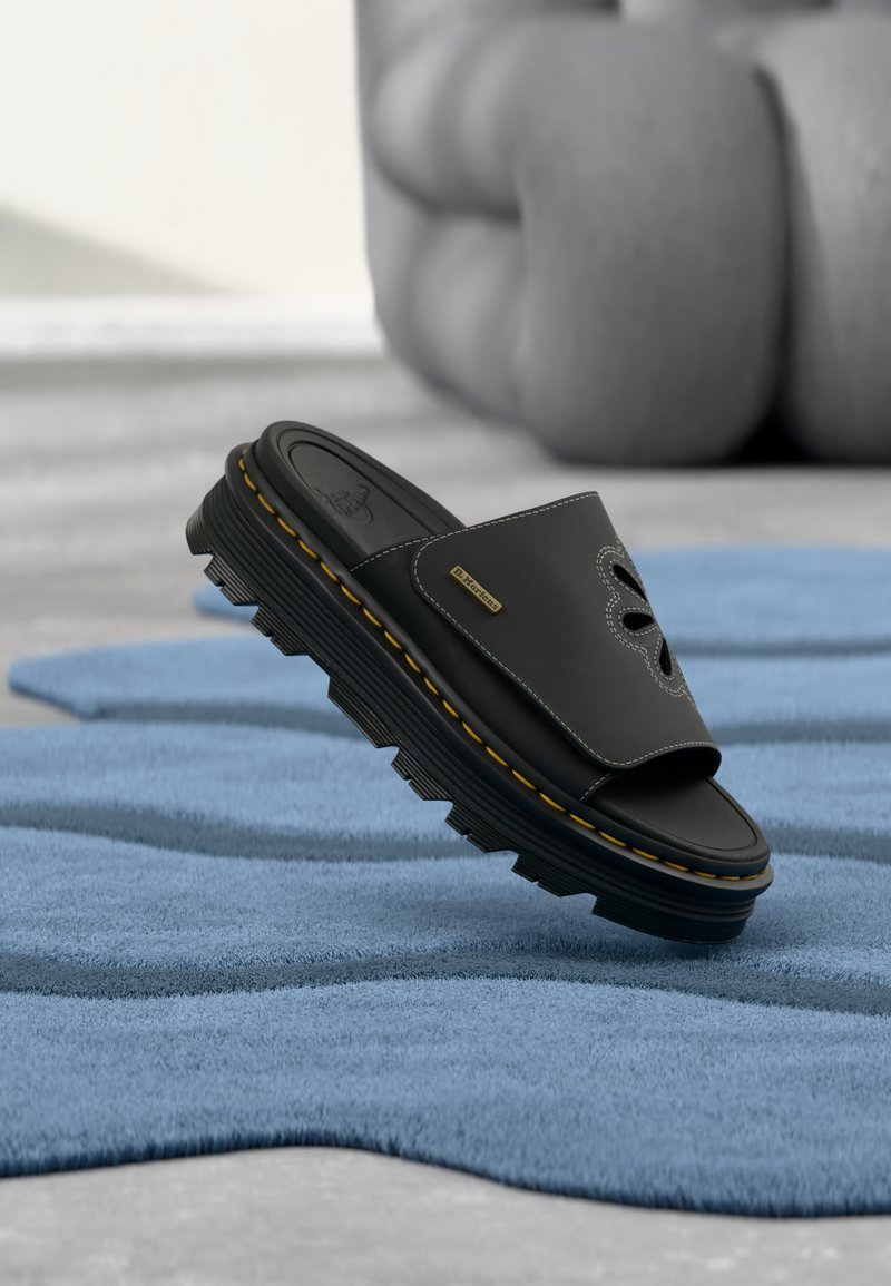 Μαύρο δερμάτινο σανδάλι slide Dr. Martens με κίτρινη ραφή σε παχύ, ανάγλυφο πάτο, τοποθετημένο σε μπλε υφασμάτινο χαλί με μαλακό γλυπτό στο φόντο.