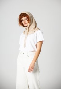 Beżowy dzianinowy szalik z kapturem o gładkiej fakturze i regulowanymi sznurkami, noszony na białej koszulce i jasnych spodniach.