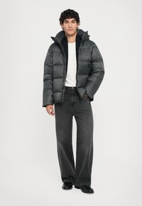 Dunkelgraue Pufferjacke mit Kapuze, gestepptem Design und Reißverschluss, kombiniert mit locker sitzenden schwarzen Jeans und schwarzen Schuhen.