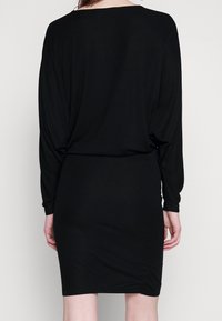 Czarna sukienka z długimi, zwisającymi rękawami, szerokim dekoltem w łódkę i dopasowaną sylwetką, z delikatnym marszczeniem w talii.