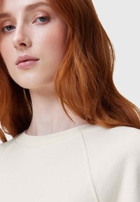 Suéter crema de punto con cuello redondo y ribetes acanalados. Textura suave, con un diseño minimalista y un sutil logotipo en el pecho.