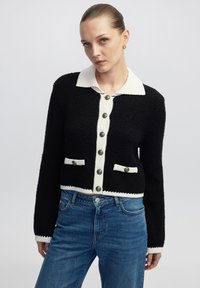Schwarze, verkürzte Strickjacke mit weißem Kragen, strukturiertem Stoff, kontrastierenden Taschendetails und großen grauen Knöpfen auf der Vorderseite.
