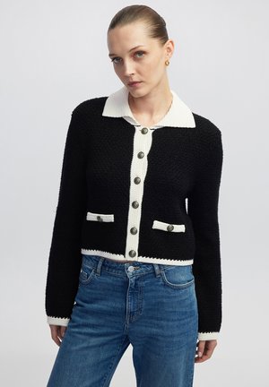 Cárdigan corto negro con un cuello blanco, tejido texturizado, detalles de bolsillos en contraste y grandes botones grises en la parte delantera.