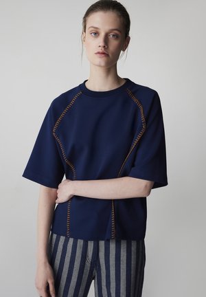 Młoda kobieta o jasnej karnacji i niebieskich oczach, ubrana w granatową bluzkę z pomarańczowymi przeszyciami oraz szaro-niebieskie spodnie w pionowe paski, stojąca.