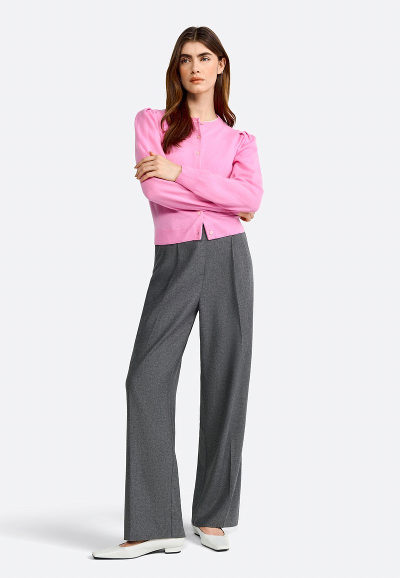 Cardigan rose à côtes avec des manches bouffantes, associé à un pantalon large gris et des chaussures plates blanches. Boutons visibles sur le cardigan.