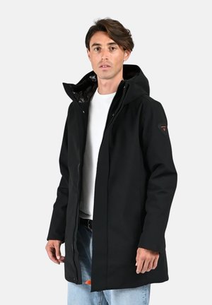 Homme portant un parka à capuche noir, une chemise blanche et un jean bleu clair, debout devant un fond uni gris clair.