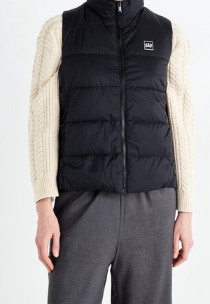 Zwart doorgestikt mouwloos vest met ritssluiting aan de voorkant en klein wit GAP-logo, gedragen over een crèmekleurige kabelgebreide trui en grijze broek.