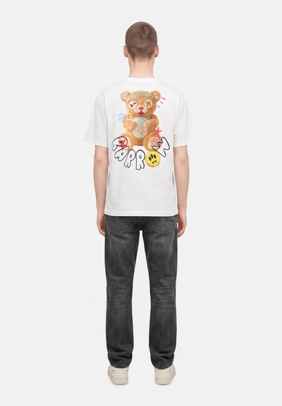 Personne portant un T-shirt blanc avec un ours en peluche coloré et le texte "Barrow" dans le dos, associé à un jean foncé et des baskets claires, debout de dos.