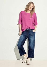 Blusa fucsia corta in tessuto testurizzato, abbinata a jeans scuri a vita alta e gamba larga e sneakers chiare, creando un look casual.