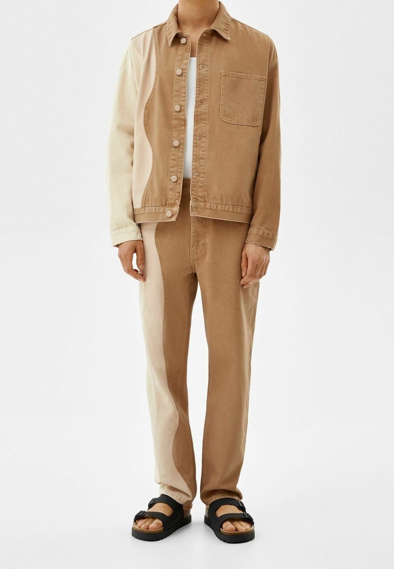 Personne portant une veste et un pantalon en denim bicolores beige et marron, associés à des sandales noires, debout sur fond blanc.