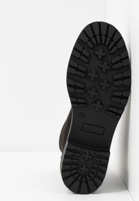 Semelle en caoutchouc noir à profil profond, présentant un motif texturé. Le corps de la chaussure est en tissu foncé, possiblement en daim, avec un design épuré.