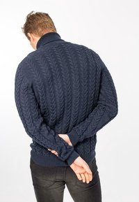 Mörkblå stickad tröja med kabelmönster, med hög krage och ribbade ärmslut. Modell visas bakifrån med händerna hopknäppta.