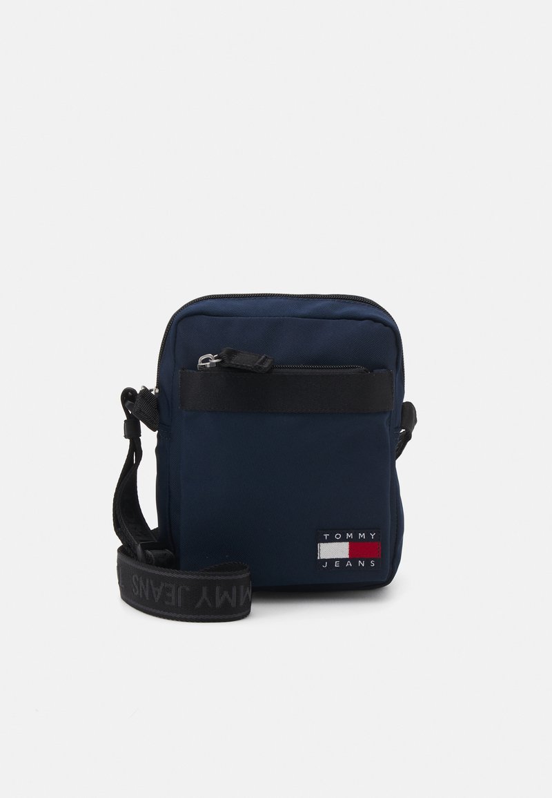 Borsa a tracolla blu marina realizzata in tessuto resistente, dotata di chiusura con zip, tracolla regolabile e un patch del logo con accenti rosso, bianco e blu.