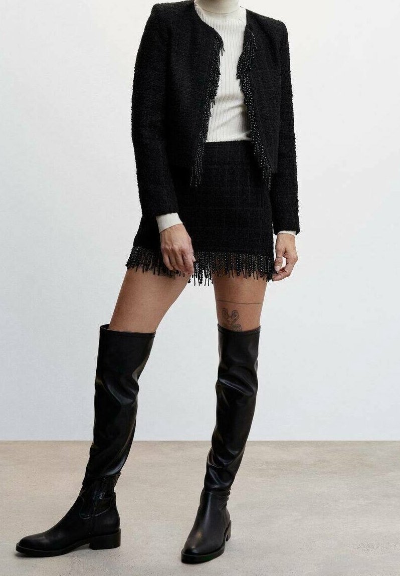 Femme portant une veste courte noire à franges, une mini-jupe à franges assortie, un haut blanc côtelé et des bottes en cuir noires montantes jusqu'aux cuisses.