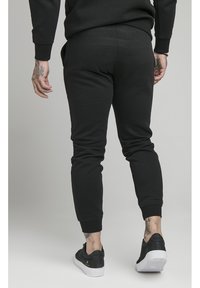 Černé teplákové kalhoty s zužujícím se střihem, žebrovanými lemy a hladkou tkaninou, kombinované s černými teniskami a viditelnými tetováními na zápěstí.