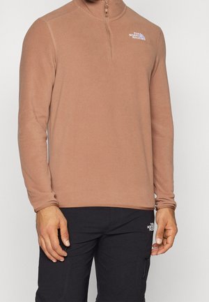 Brauner Fleece-Pullover mit halbseitigem Reißverschluss-Kragen. Enthält ein kleines weißes Logo auf der linken Brust und lange Ärmel. Getragen mit dunklen Hosen.