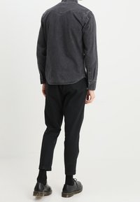 Camicia in denim grigio scuro con colletto classico, dettaglio a mantellina sul retro e polsini con bottoni, abbinata a pantaloni neri a gamba stretta e scarpe con lacci.