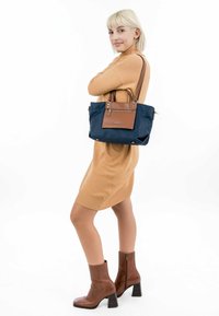 Borsa a tracolla blu navy con un accento in pelle marrone, due manici superiori e una tasca frontale con zip. Tessuto texturizzato con dettagli in metallo argentato.