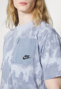 Light blue short-sleeve t-shirt with a dotted pattern, features a zippered chest pocket, and black Nike logo on the pocket.
