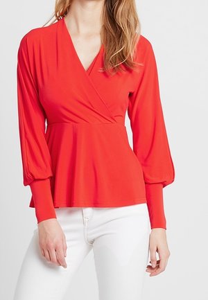 Femme portant un chemisier rouge à manches longues, style cache-cœur, avec des poignets plissés et un pantalon blanc, visible du cou aux hanches.