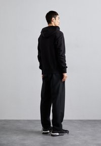 Felpa nera con cappuccio, vestibilità ampia, abbinata a pantaloni scuri e larghi. L'outfit è completato da sneakers nere con accenti bianchi e gialli.