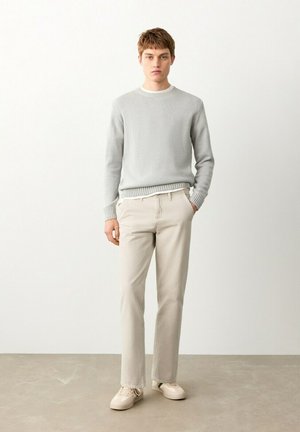 Young man standing, wearing light grey sweater, beige pants, and off-white sneakers, against plain white wall and grey floor.