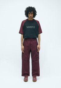 Schwarzes und burgunderfarbenes T-Shirt mit Timberland-Logo, kombiniert mit weit geschnittenen burgunderfarbenen Hosen. Braune Schnürschuhe runden das Outfit ab.