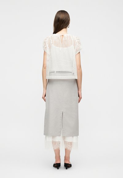 Femme debout, de dos, portant un haut blanc à manches courtes à mailles ouvertes, une jupe midi grise claire fendue au dos, une sous-couche en dentelle blanche, et des talons noirs.